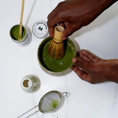 MatchaMatcha UK Ceremonial Grade matcha being prepared with a bamboo whisk in a matcha bowls. Shows tin of MatchaMatcha UK ceremonial grade matcha.